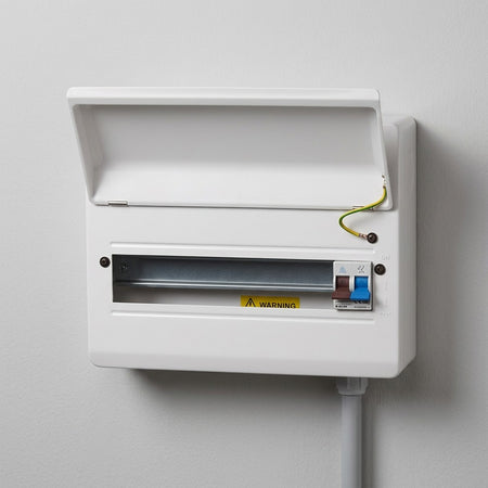 A white electrical distribution board or switchbox mounted on a light gray wall, with the lid open, showing a metal busbar, a yellow warning label, and a small blue-and-white switch.