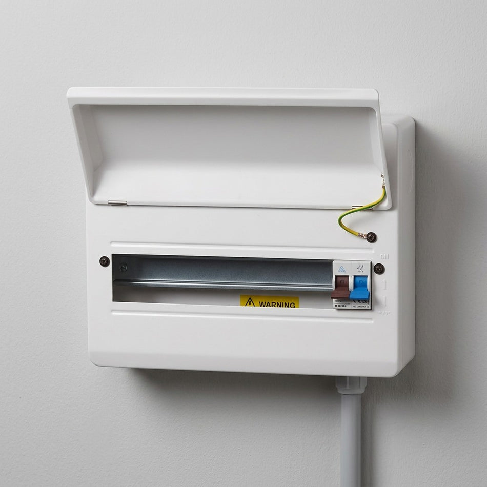 A white electrical distribution board or switchbox mounted on a light gray wall, with the lid open, showing a metal busbar, a yellow warning label, and a small blue-and-white switch.