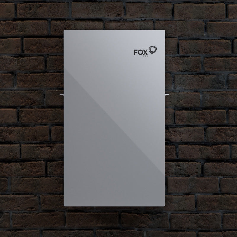 A silver rectangular wall-mounted solar battery storage system by FOX ESS, shown against a dark brown brick wall background.