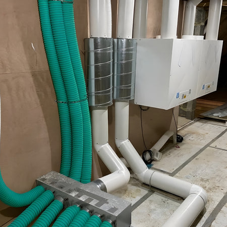 Industrial ventilation system with white ductwork, galvanized steel ducts, and bright green flexible supply hoses on a concrete floor against a cardboard wall.