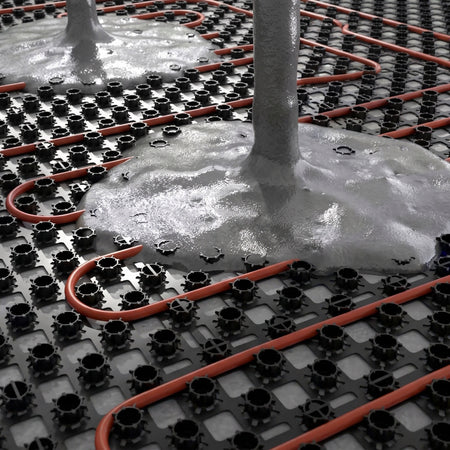 Close-up of a radiant floor heating system showing orange heating pipes laid over a black grid base, with wet concrete or foam insulation around the pipes and a vertical pipe column.