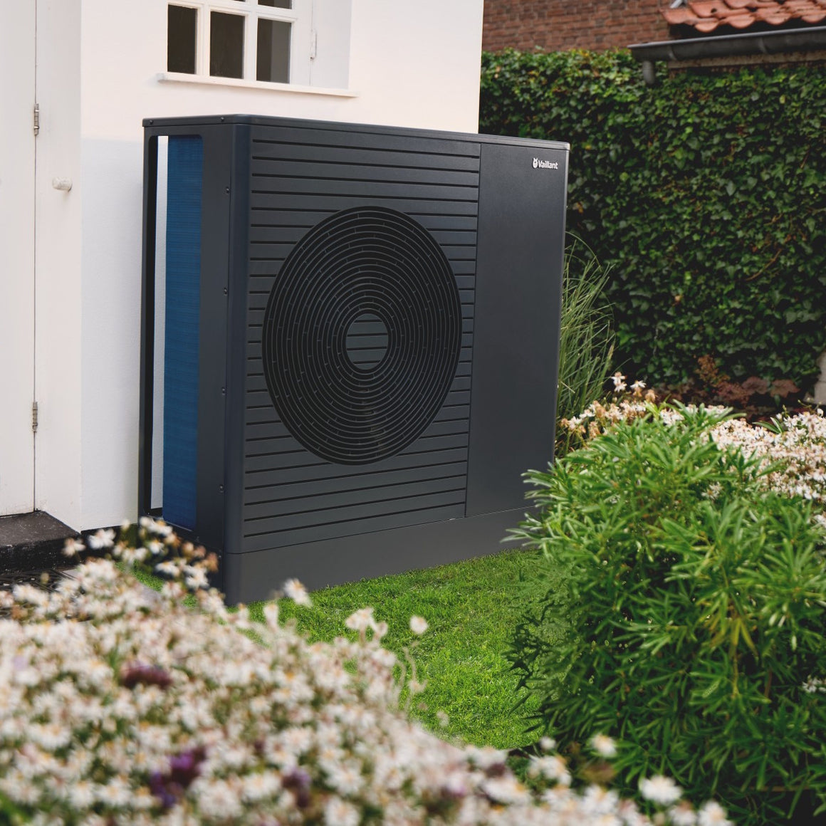 A dark gray outdoor heat pump or air source heat system unit standing on grass in front of a white house, with hedges and flowering plants around it. The brand name 'EcoFlow' is visible on the upper right corner of the unit.