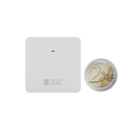 A square white Delta Dore electronic device with a small central slit and the Delta Dore logo near the bottom, shown next to a 2 Euro coin for scale on a plain white background.